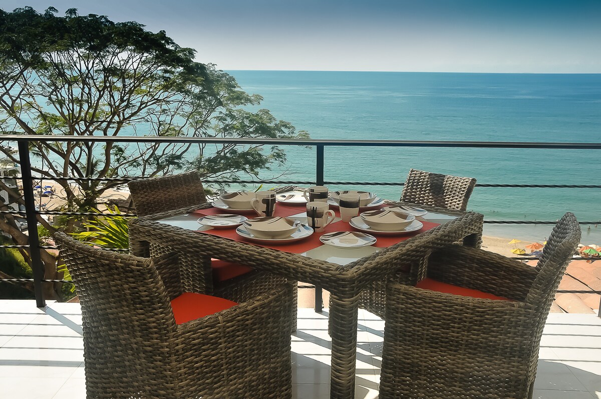 An outdoor dining area is set with a square table surrounded by four woven chairs featuring orange cushions. Ocean views are visible in the background, complemented by lush greenery. A selection of dishes and utensils is arranged on the table, inviting guests to enjoy meals in a scenic setting.