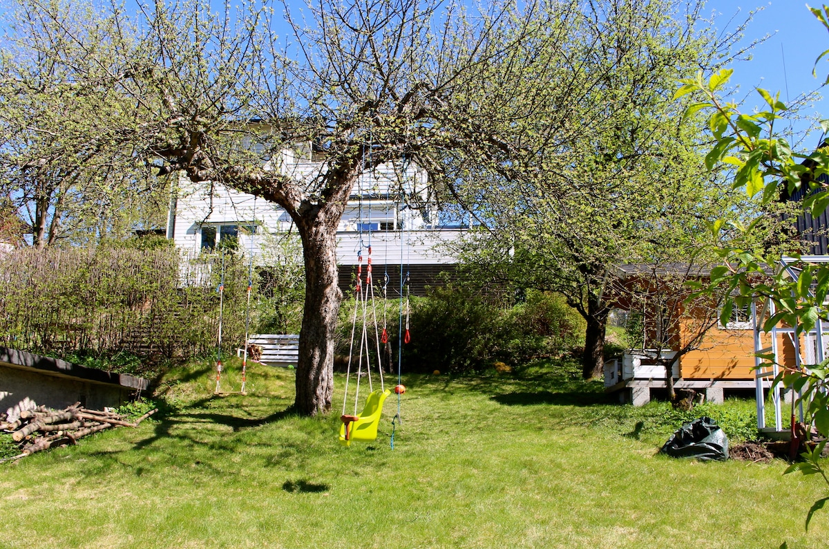 A spacious backyard is depicted, featuring a large tree with budding leaves, a green lawn, and a play area with swings. A playhouse is visible in the background, surrounded by fruit trees and a tidy garden, creating an inviting outdoor space.