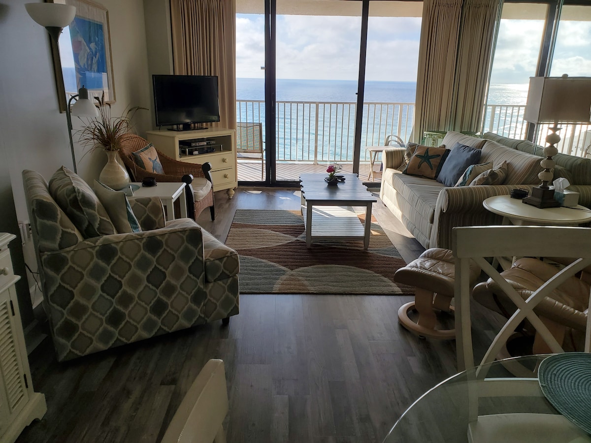 A comfortable living area features large sliding glass doors offering a view of the Gulf. Soft furnishings, including a light-colored sofa and patterned chairs, create a cozy atmosphere. A coffee table sits centrally on a patterned rug, while a flat-screen TV is placed on a cabinet nearby.