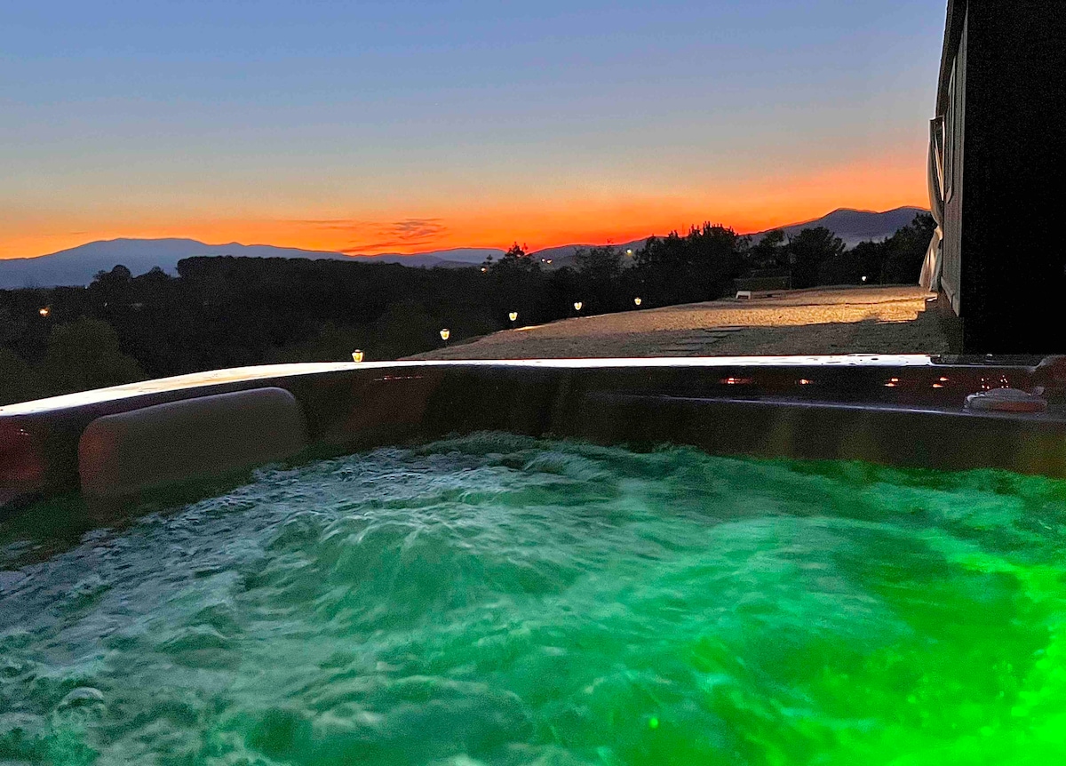 A hot tub is positioned on a deck, featuring illuminated green water, with a stunning sunset casting warm hues over a mountain view in the background. Soft outdoor lighting adds to the serene ambiance, creating a calm space for relaxation.