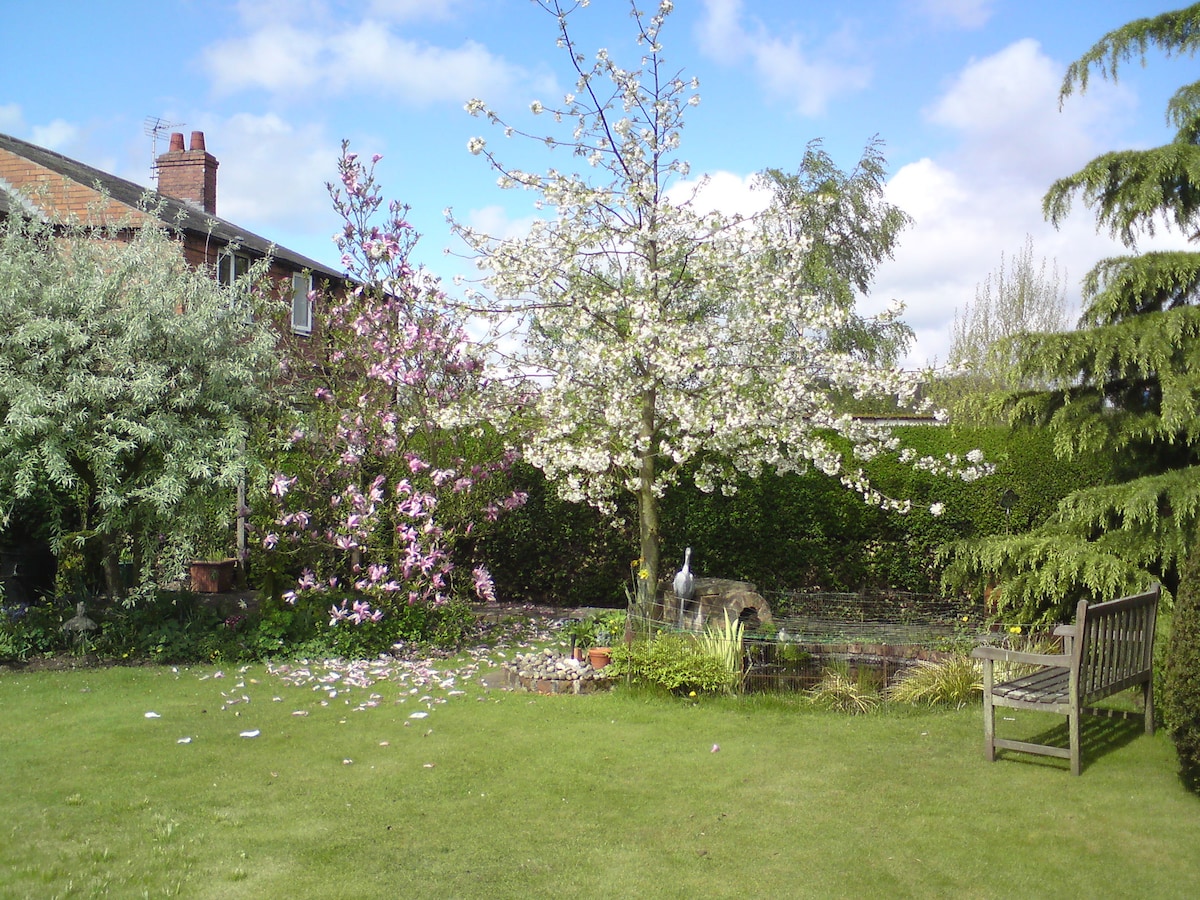 A peaceful garden scene is presented, featuring a variety of flowering trees including pink and white blossoms. Lush green grass covers the ground, with a small pond visible among the foliage. A wooden bench is placed near the water, offering a serene spot to relax.