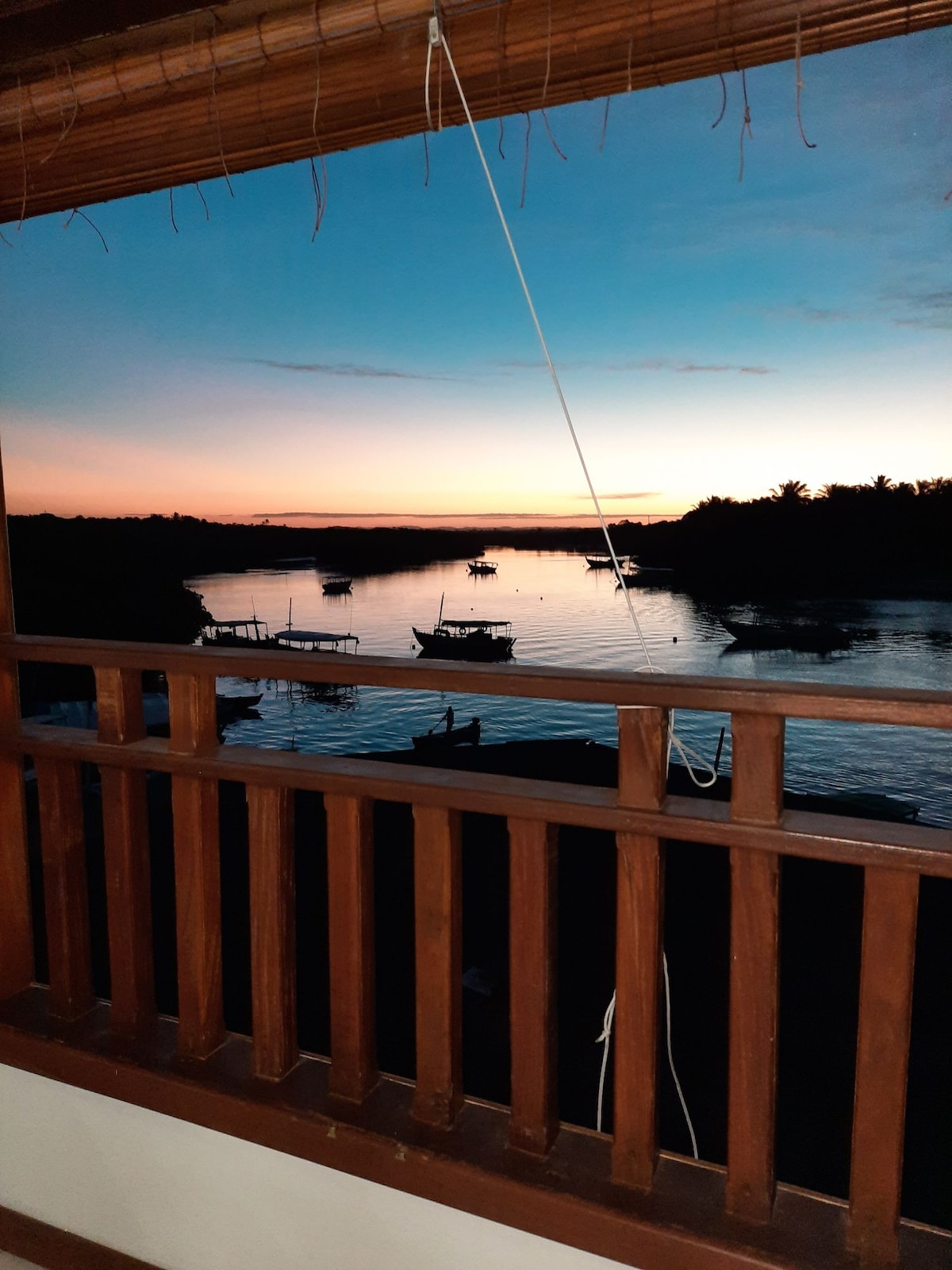 A serene view from a wooden balcony showcases a calm waterway at dusk, with silhouettes of boats gently floating. The horizon is painted in soft hues of orange and blue, providing a tranquil backdrop to the natural landscape.
