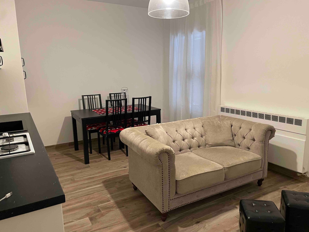 A comfortable living space features a tufted beige sofa positioned in front of a black dining table surrounded by six chairs. Natural light enters through sheer curtains, enhancing the warm wooden floor and creating a welcoming atmosphere.