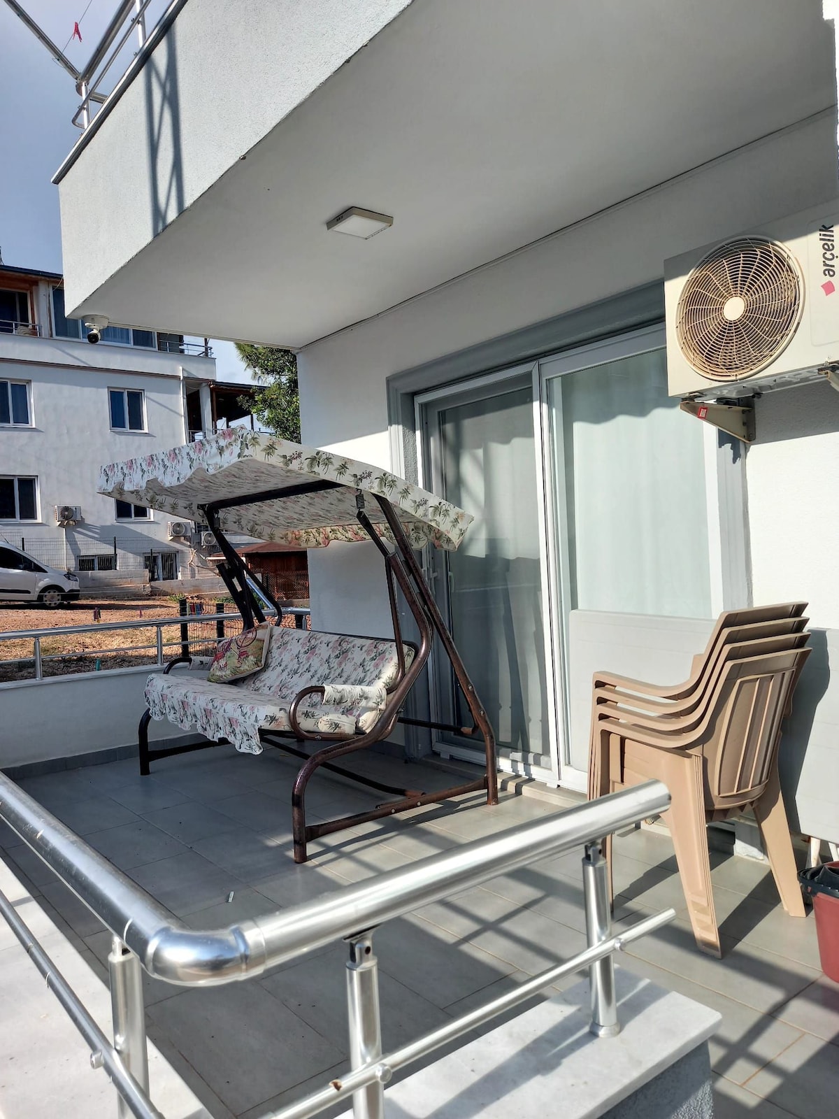 A shaded outdoor area is presented, featuring a swing seat with floral upholstery. Nearby, a set of four stacked chairs rests against a wall. An air conditioning unit is mounted above the glass doors leading into the interior space.
