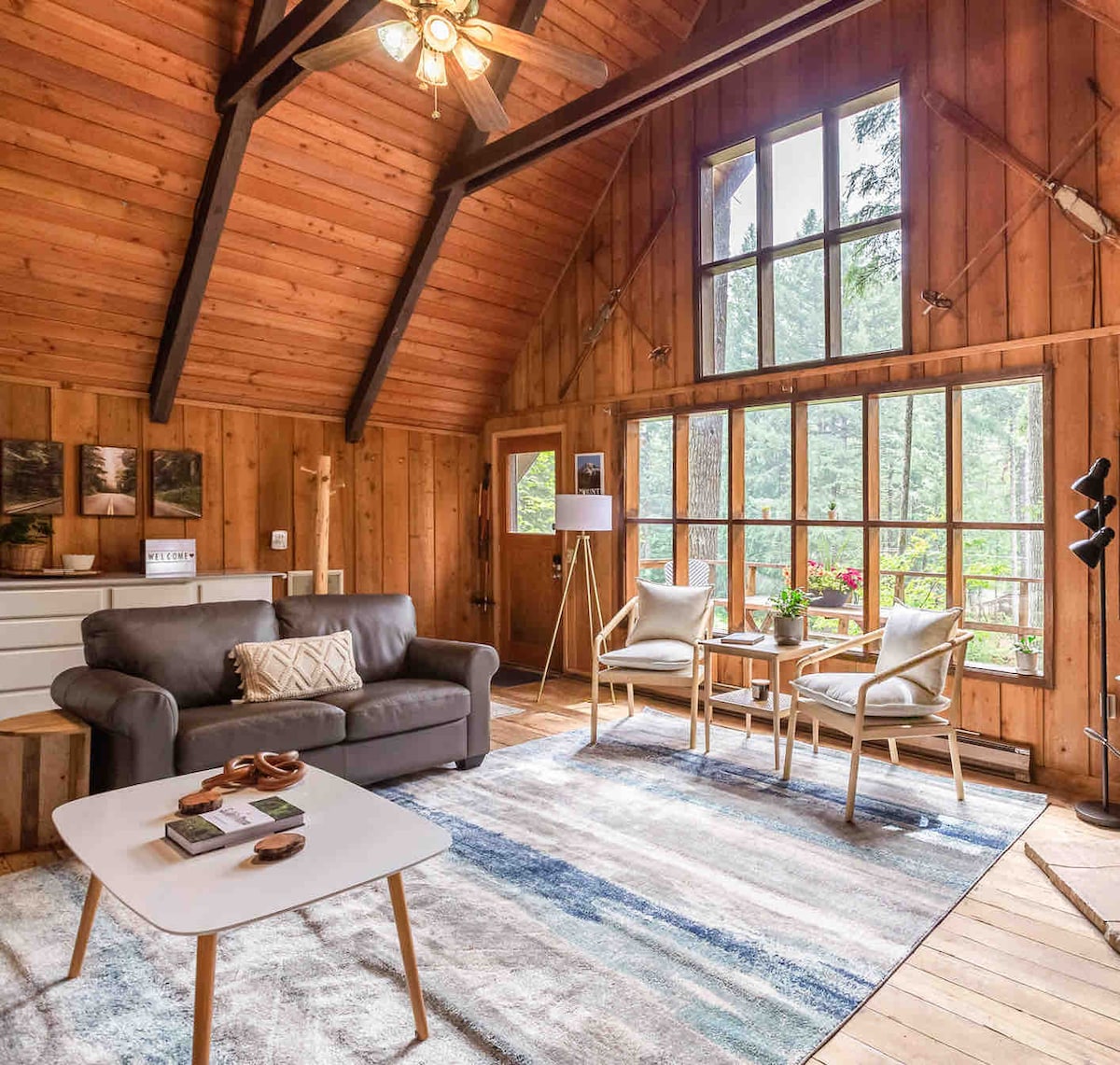 A spacious living area features high ceilings and warm wood paneling, illuminated by large windows that offer views of surrounding greenery. A comfortable sofa is positioned next to two chairs, set on a soft area rug. A coffee table showcases books and decor.