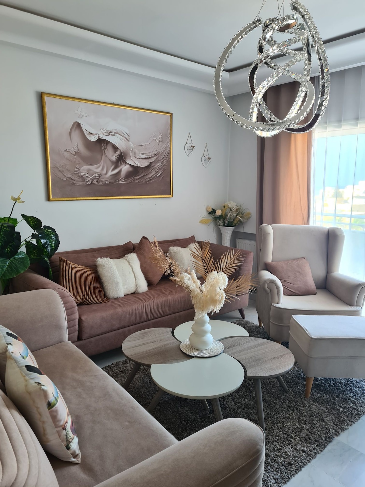A modern living area features a comfortable sofa and armchair arrangement, complemented by soft decorative pillows. A stylish chandelier provides ambient lighting, while a circular coffee table sits at the center. Plants and decorative elements add a touch of elegance, enhancing the inviting space.