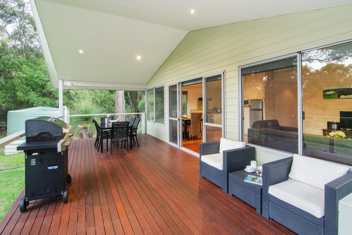 A covered timber deck features outdoor seating and dining arrangements, providing a view of the surrounding natural bush. A BBQ grill is positioned nearby, with large glass doors leading into the modern living area. Warm lighting enhances the welcoming ambiance.
