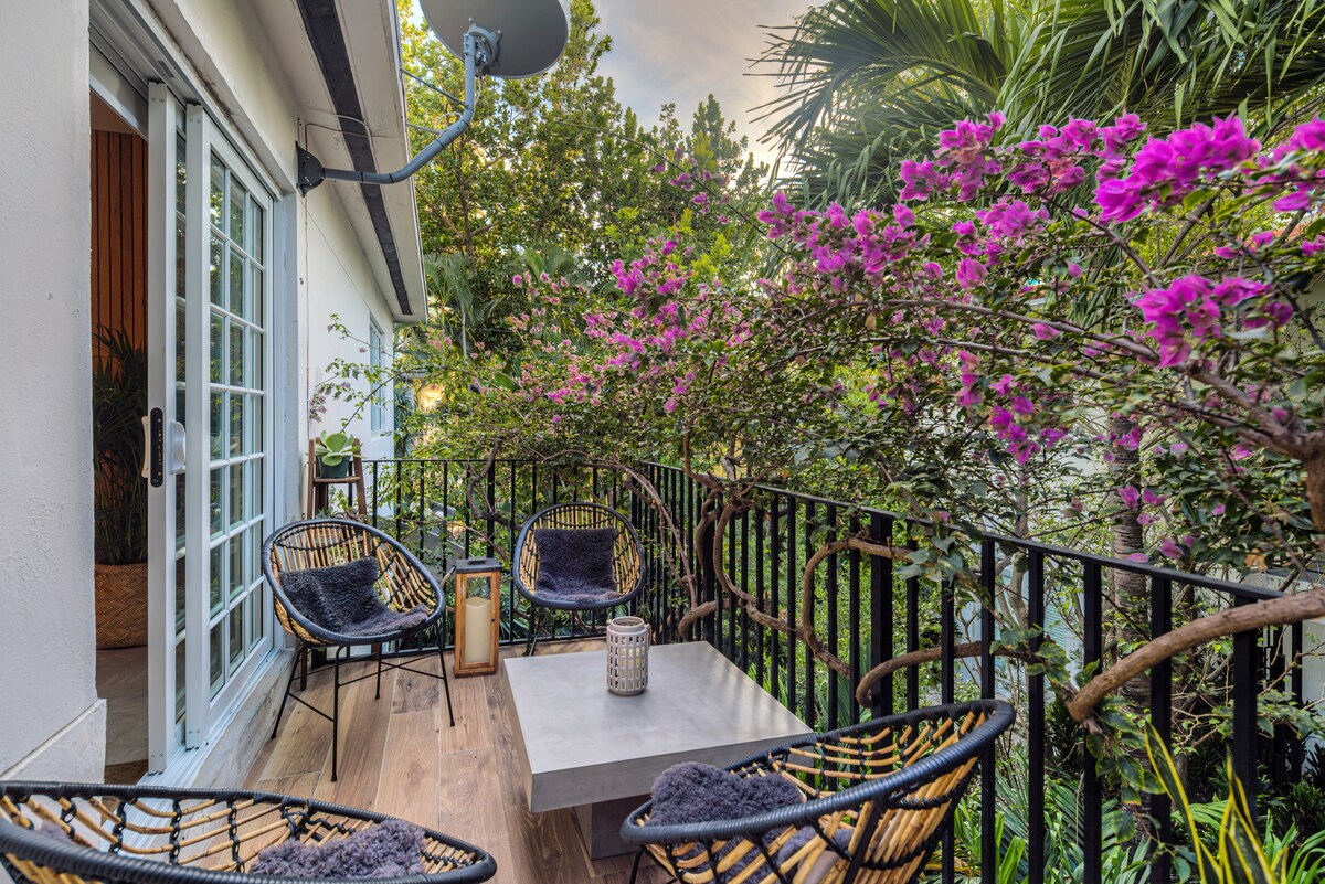 A balcony adorned with vibrant pink flowers and greenery features four woven chairs positioned around a square table. Natural wood flooring enhances the serene atmosphere, while soft cushions add comfort to the seating area. The surrounding foliage contributes to a tranquil outdoor space.