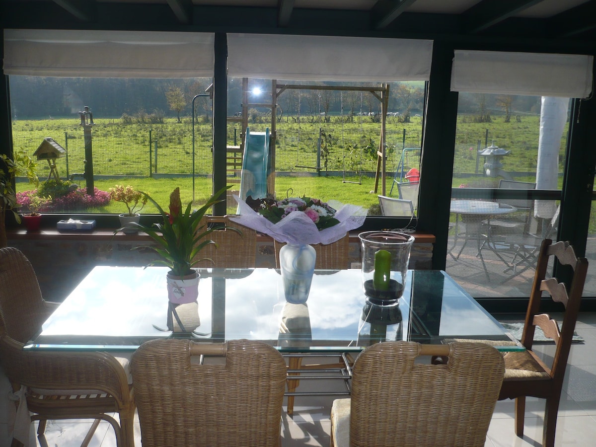 A well-lit veranda features a glass dining table surrounded by woven chairs. A bouquet rests at the center of the table, and a green field with trees is visible through the large windows. Outdoor seating and a play structure can be seen in the distance.