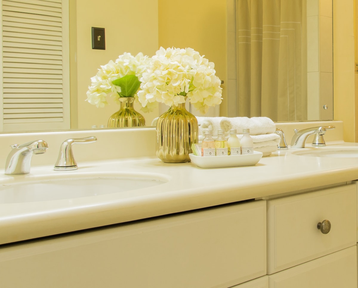 A well-appointed bathroom features a double sink with sleek chrome faucets and a neutral countertop. A decorative vase filled with white flowers adds a touch of elegance. Nearby, neatly arranged toiletries and towels are presented on a tray, providing a sense of organization.