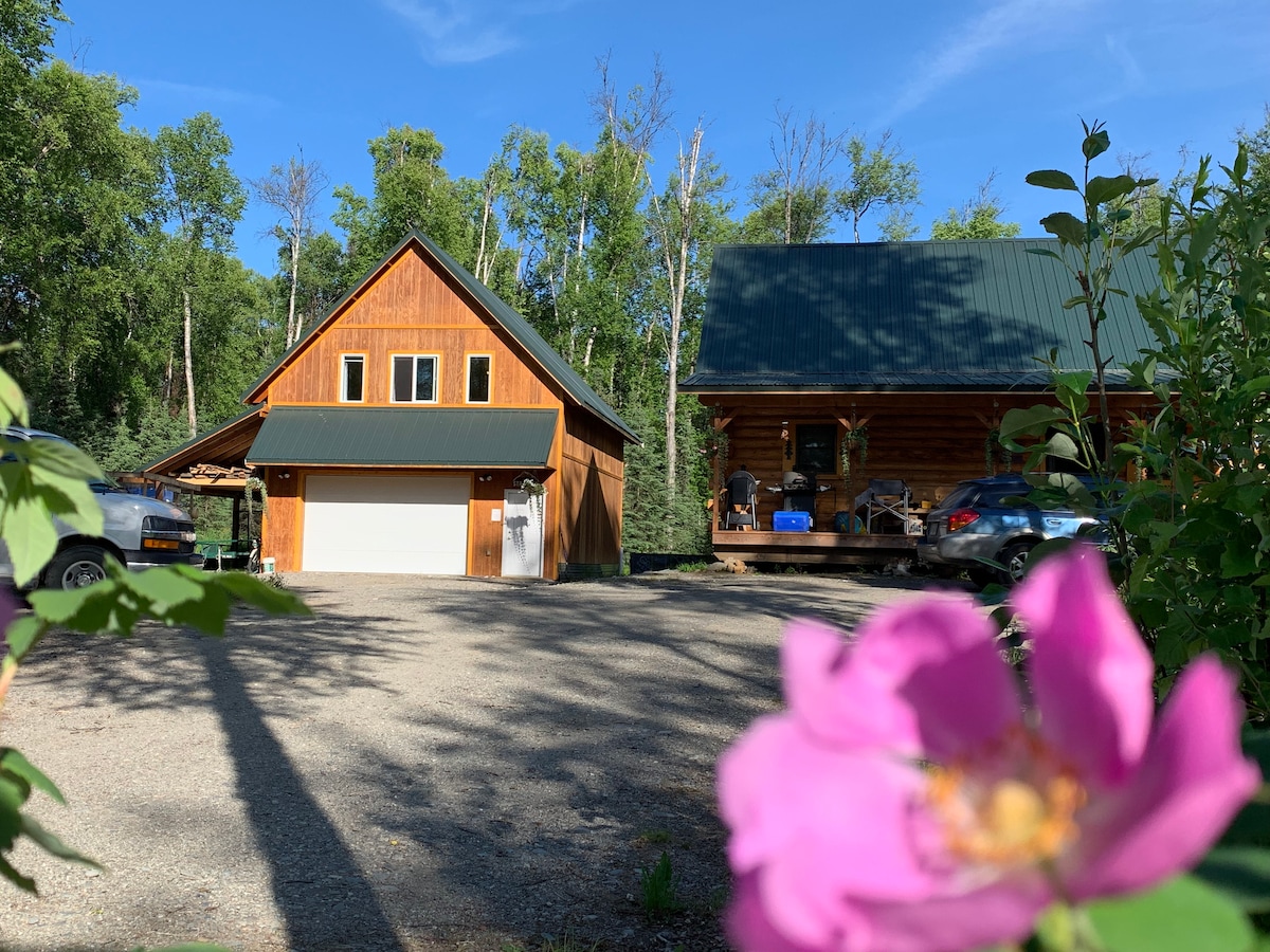 A rustic wooden structure with a green metal roof stands among trees in a natural setting. The foreground features a pink flower, while a gravel driveway leads to the garage entrance, emphasizing the property's tranquil surroundings.