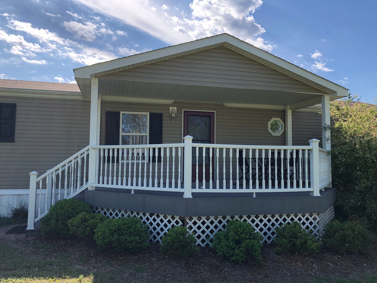The front porch features a white railing, welcoming guests with a shaded area for relaxation. Lush greenery surrounds the base, offering a sense of tranquility. The home has a neutral exterior, complemented by a simple decorative element near the entrance.