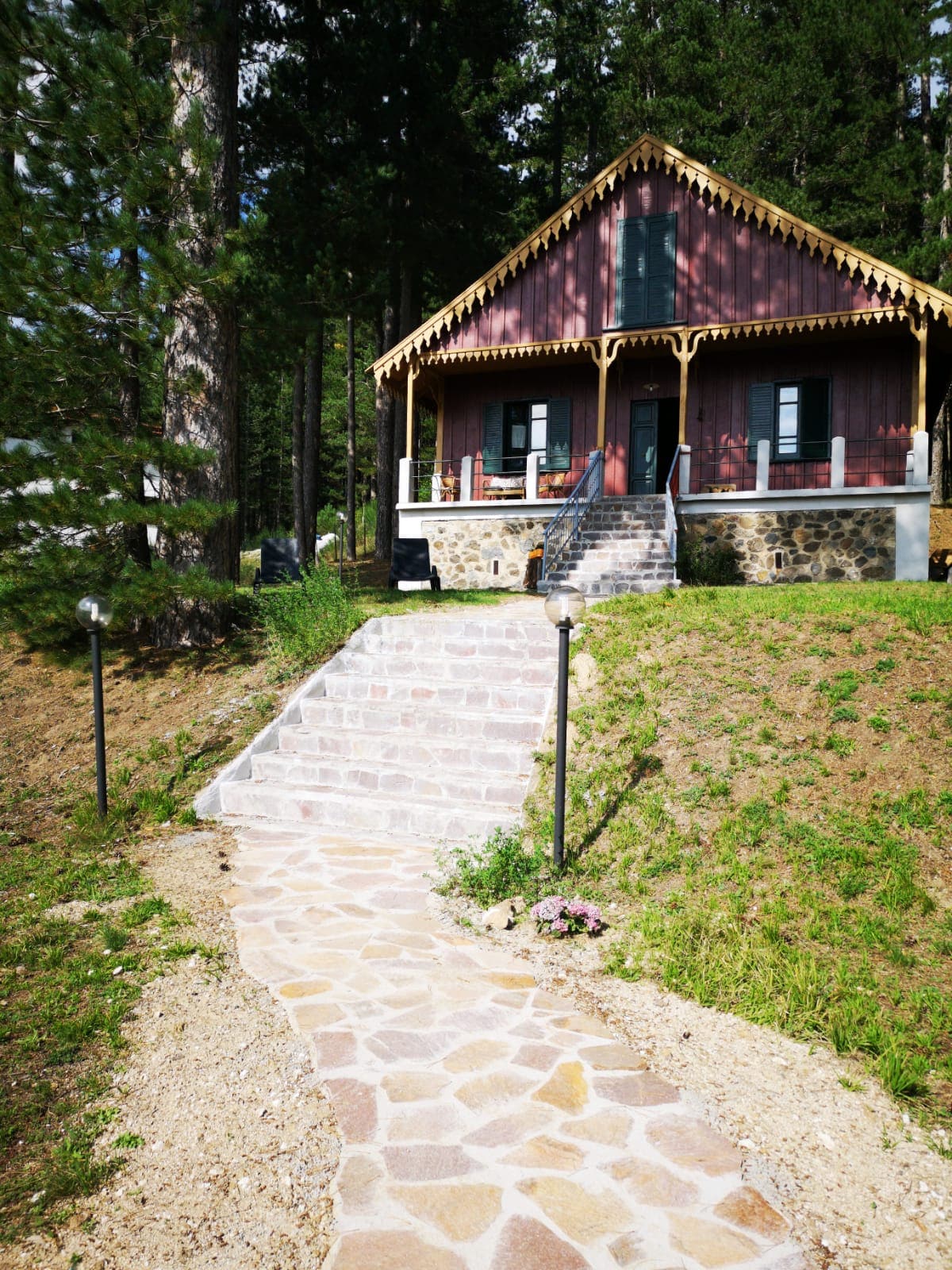A rustic cabin is situated on a gentle slope, with steps leading to a front porch adorned with intricate woodwork. Lush greenery surrounds the cabin, complimented by a pathway of natural stone. Windows are framed in dark green, providing a contrast against the warm wood exterior.