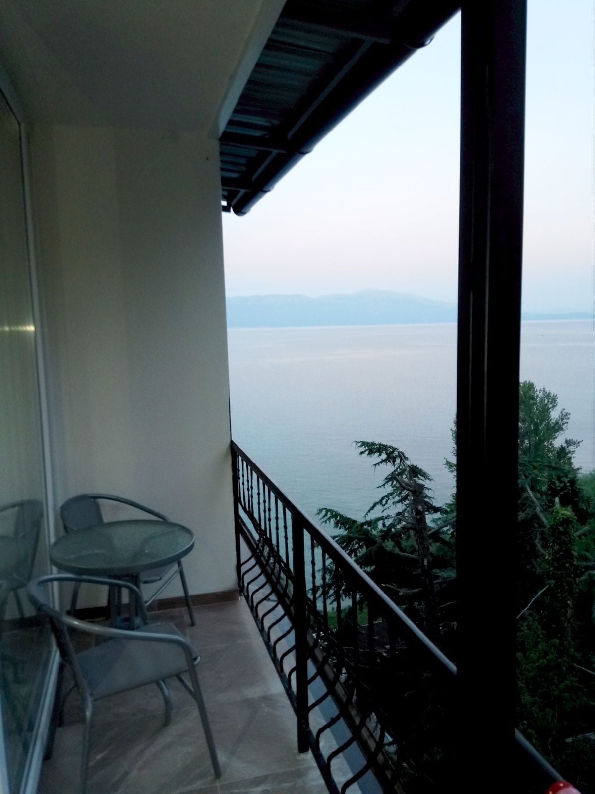 A private balcony is shown with a glass-top table and two metal chairs. The view overlooks the calm waters of the lake and distant mountains, framed by green foliage on one side, creating a serene outdoor setting.