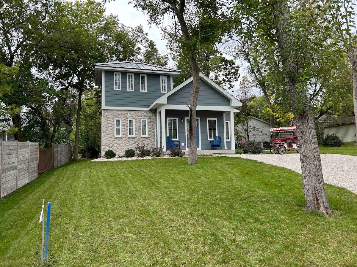 A newly constructed two-story home is framed by well-maintained grass and mature trees. The facade combines stone and siding, featuring a welcoming porch with two blue chairs. A gravel driveway leads up to the entrance, indicating a spacious outdoor area.