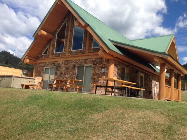 A handcrafted log and stone cabin features a green metal roof and large windows, allowing natural light to fill the space. Surrounding grassy areas include picnic tables, while a hot tub is positioned nearby, offering a serene view of the landscape.