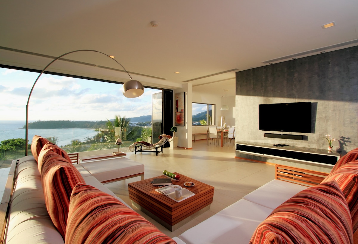 A spacious living area features a large wraparound sofa adorned with striped cushions, inviting relaxation. Floor-to-ceiling windows allow for ample natural light and an unobstructed view of the Andaman Sea. A wooden coffee table sits in the center, complemented by a sleek entertainment unit with a flat-screen TV.