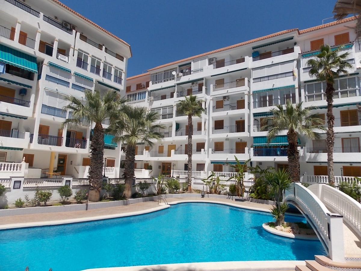 A sunlit view of the residential complex showcases a large swimming pool surrounded by palm trees. The architecture features multiple balconies and white railings, with a small bridge leading across the water, providing a relaxed and welcoming environment.