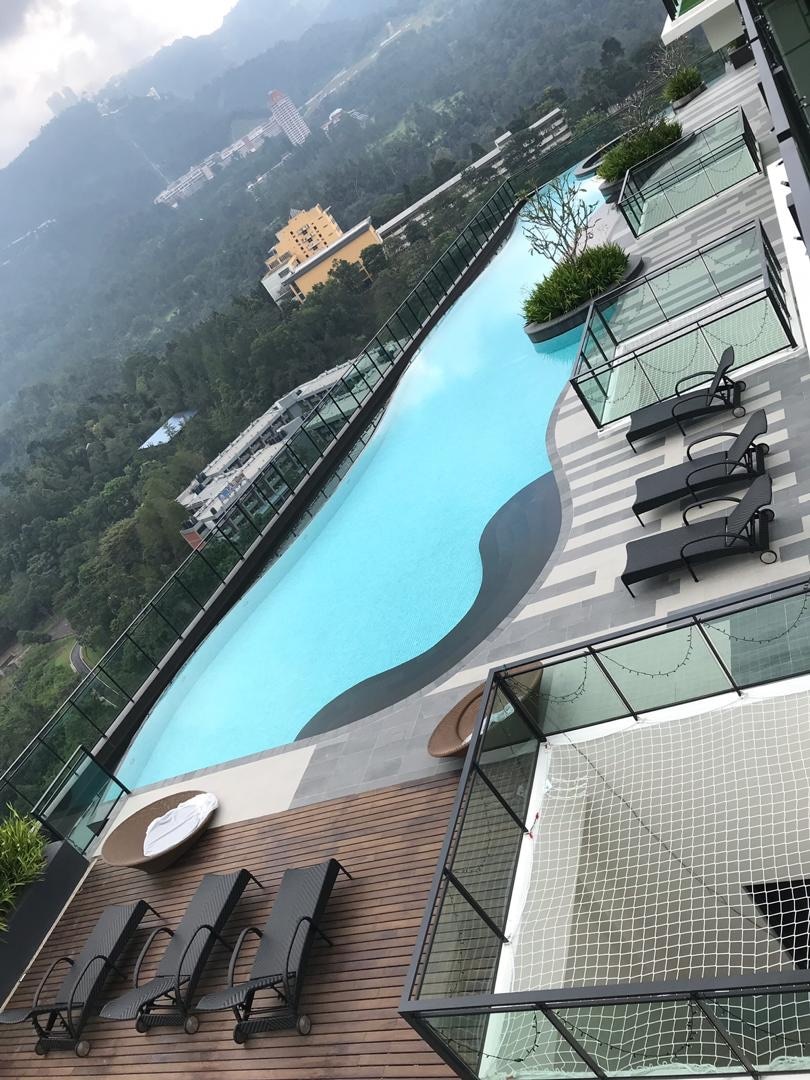 An expansive swimming pool is situated on a rooftop, featuring a smooth turquoise surface surrounded by wooden decking. Lounge chairs are arranged along the pool's edge, with views of the surrounding mountains and lush greenery. Balconies with glass railings overlook the scene.