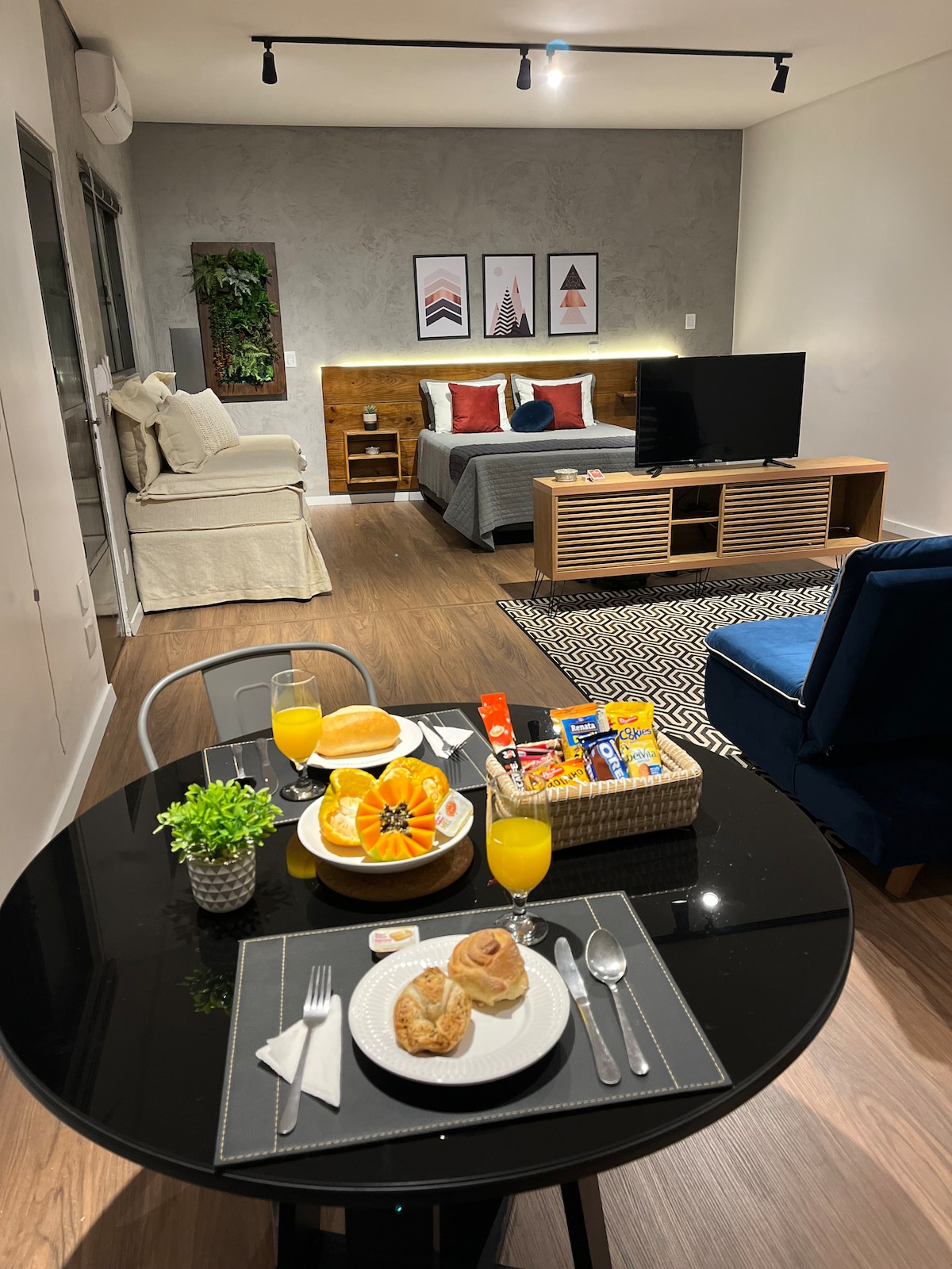 A dining area is set with a black table, showcasing plates of pastries and drinks. In the background, a cozy sleeping area features a gray bed with accent pillows, a light gray sofa, and a television on a wooden console. Soft lighting enhances the welcoming ambiance.