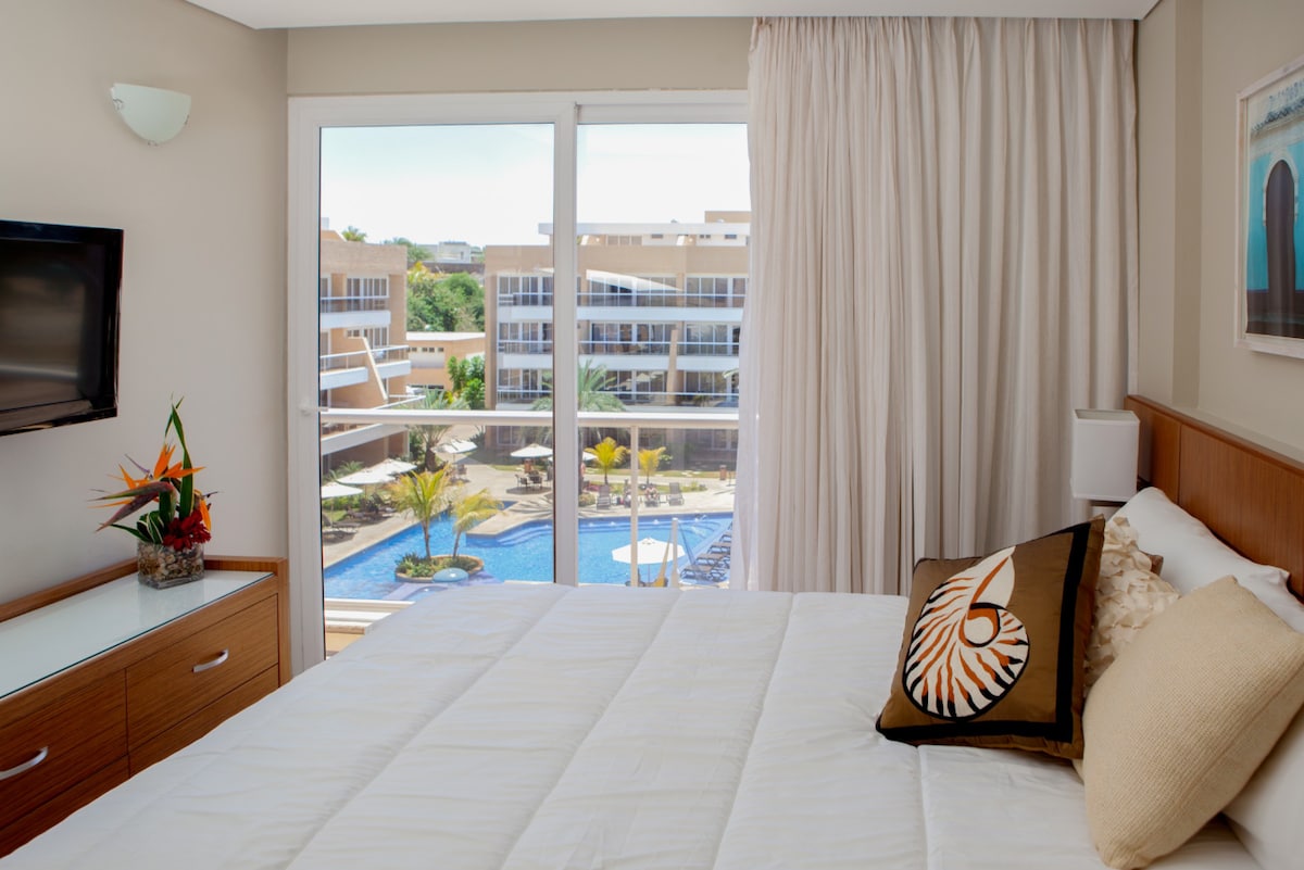 A well-appointed bedroom features a spacious bed with light bedding, accentuated by a decorative pillow. Natural light enters through large glass doors, providing views of the outdoor pool area and palm trees beyond. A flat-screen TV is mounted on the wall, adding modern convenience.