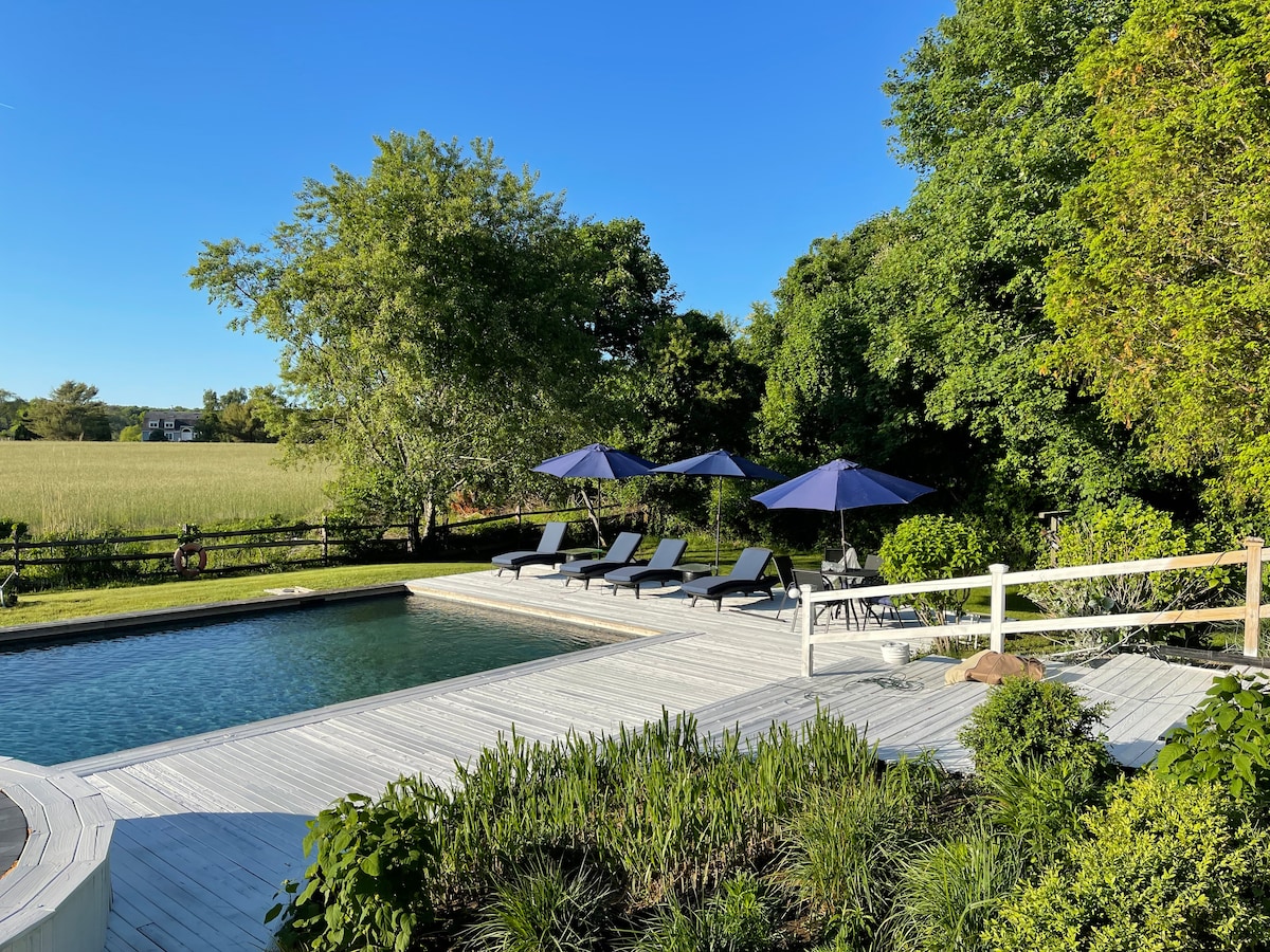 A saltwater pool is surrounded by a wooden deck and lush landscaping. Lounge chairs with umbrellas are arranged on one side, offering shaded seating. In the background, a green field is visible, contributing to the serene environment.