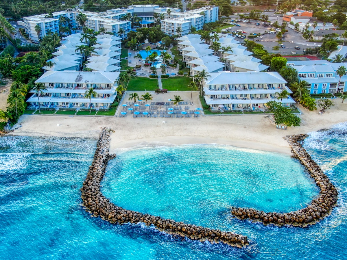 An aerial view captures the resort-style complex, showcasing multiple buildings arranged around a sandy beach. A rock barrier lines the shoreline, providing a protective cove. The turquoise waters gently lap against the shore, while sun loungers are positioned for relaxation amidst tropical landscaping.