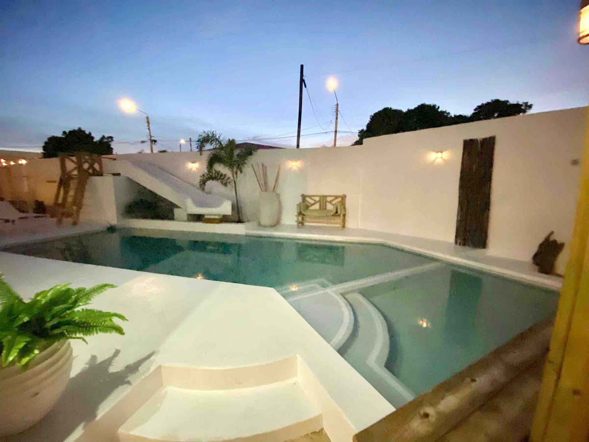 A serene outdoor space features a private pool surrounded by white walls, complemented by a sliding water feature. Soft lighting from wall sconces creates a calm ambiance, while a small seating area is positioned near the water. Lush greenery is present in planters.