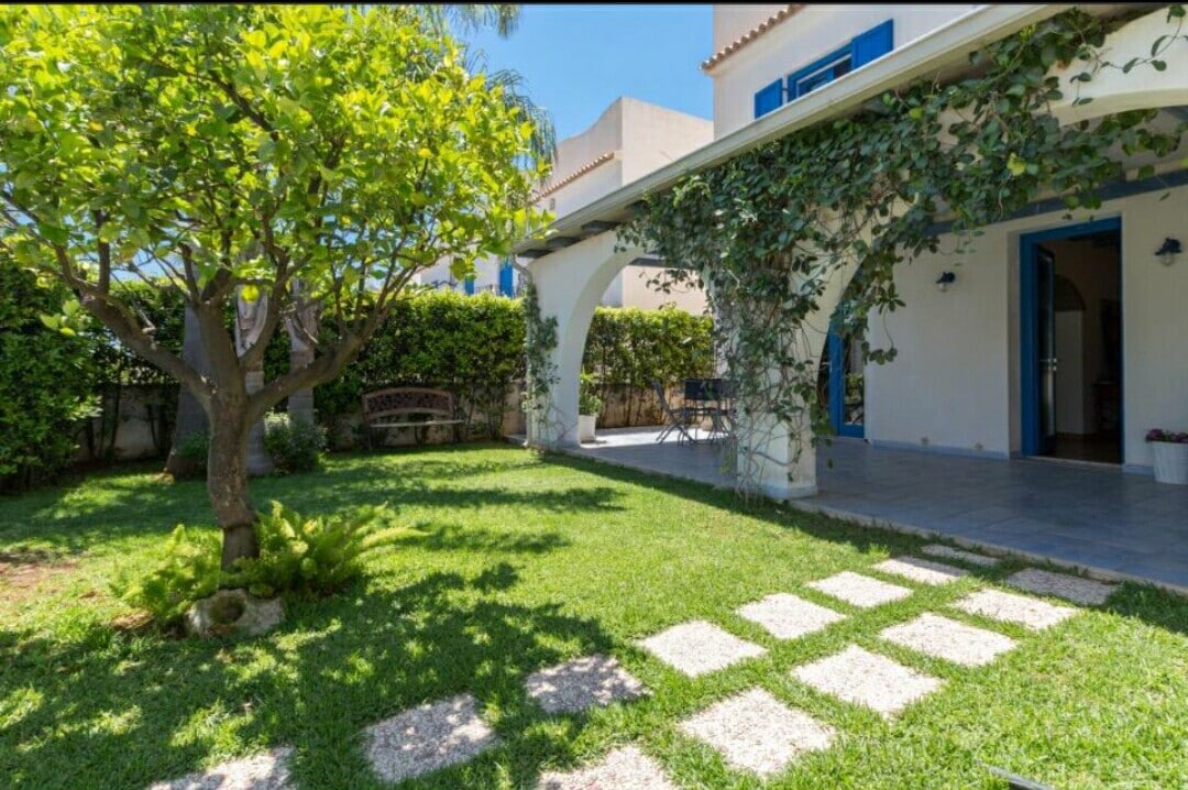 A private garden is featured, showcasing a lush green lawn and shaded areas from the leafy tree. Elegant stone pathways lead to a shaded veranda, which is furnished with seating for outdoor relaxation.