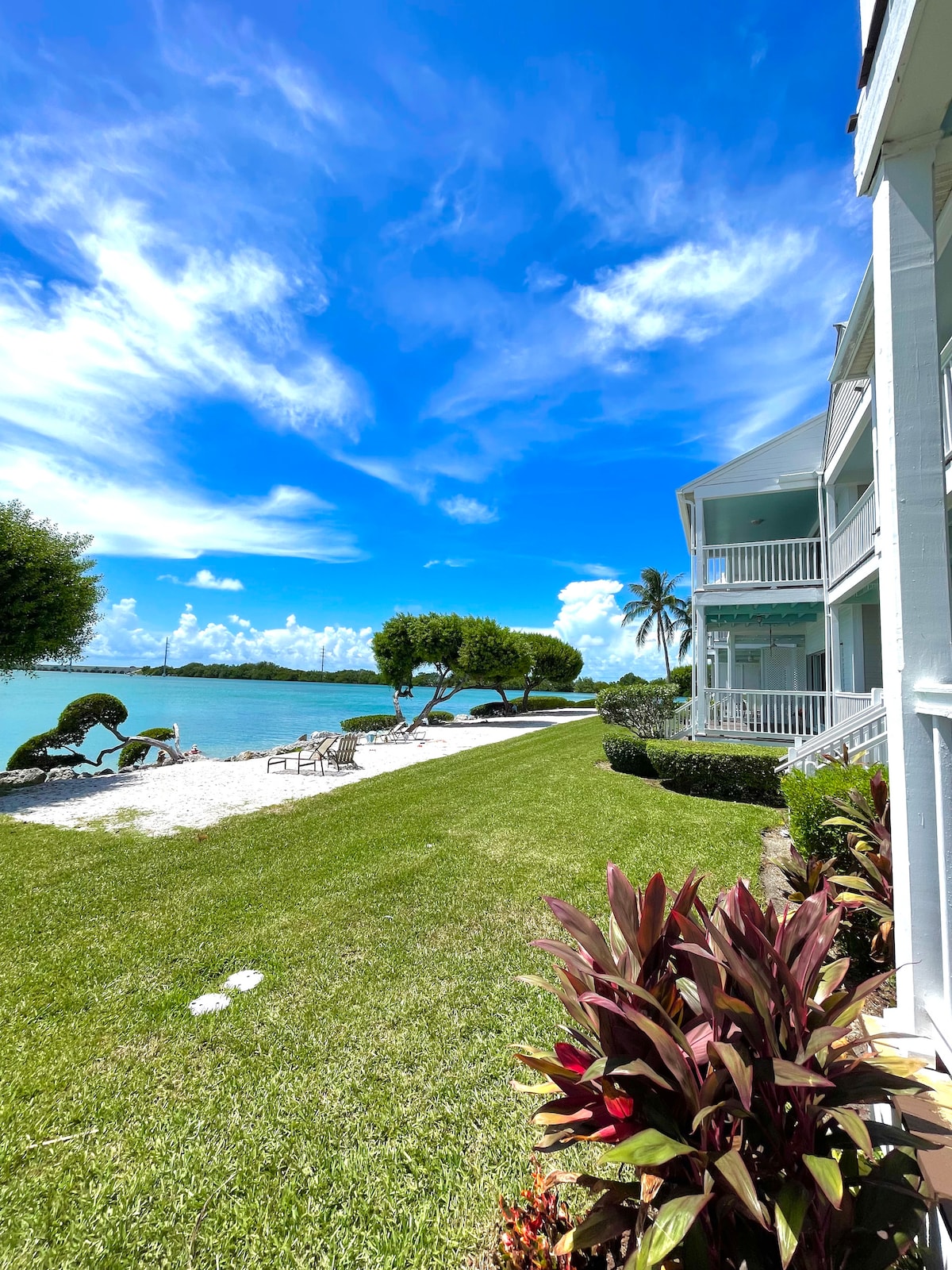 The image captures a serene beachfront view featuring neatly manicured grass leading to a sandy shore. Vibrant foliage lines the foreground, while the calm water reflects a bright blue sky with scattered clouds. Nearby, the villa stands with white exterior and balconies overlooking the water.