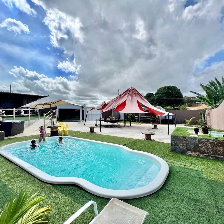 A refreshing pool is surrounded by vibrant green grass, with a large tent providing shaded seating. Children are seen enjoying the water, while stylish lounge chairs and additional outdoor features complement the space. A backdrop of fluffy clouds adds to the sunny ambiance.