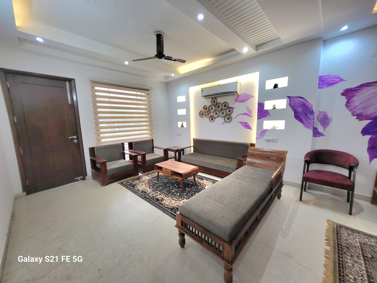 A spacious living room is presented, featuring comfortable seating arrangements with wooden furniture and soft gray cushions. A central wooden coffee table anchors the space. Decorative wall art and ambient lighting enhance the modern decor, while natural light filters through the window treatments.