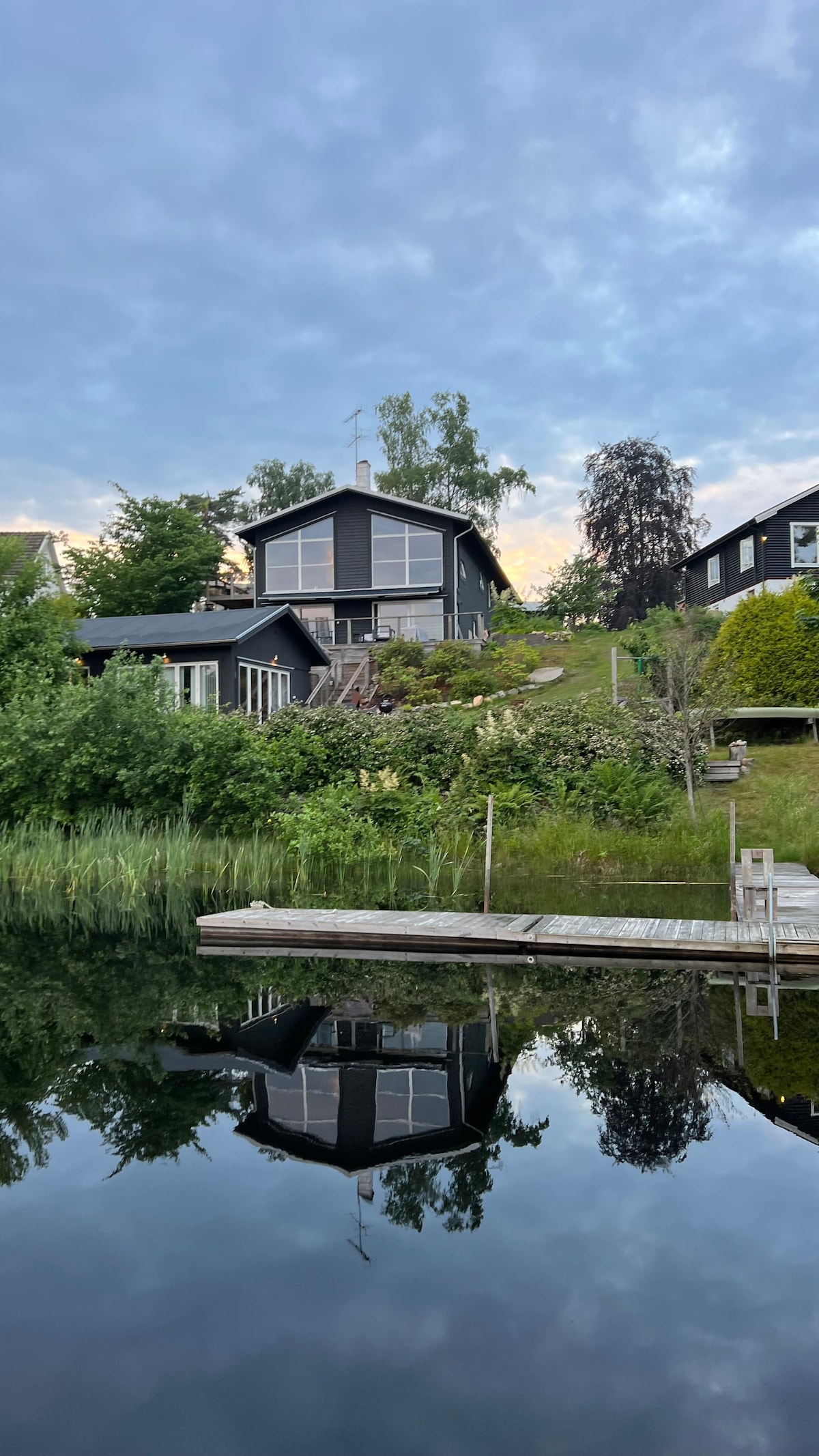 A serene lakeside cabin is reflected on the calm water, surrounded by lush greenery. The two-story structure features large windows and a welcoming staircase leading up to the entrance, creating a harmonious blend with the natural landscape.
