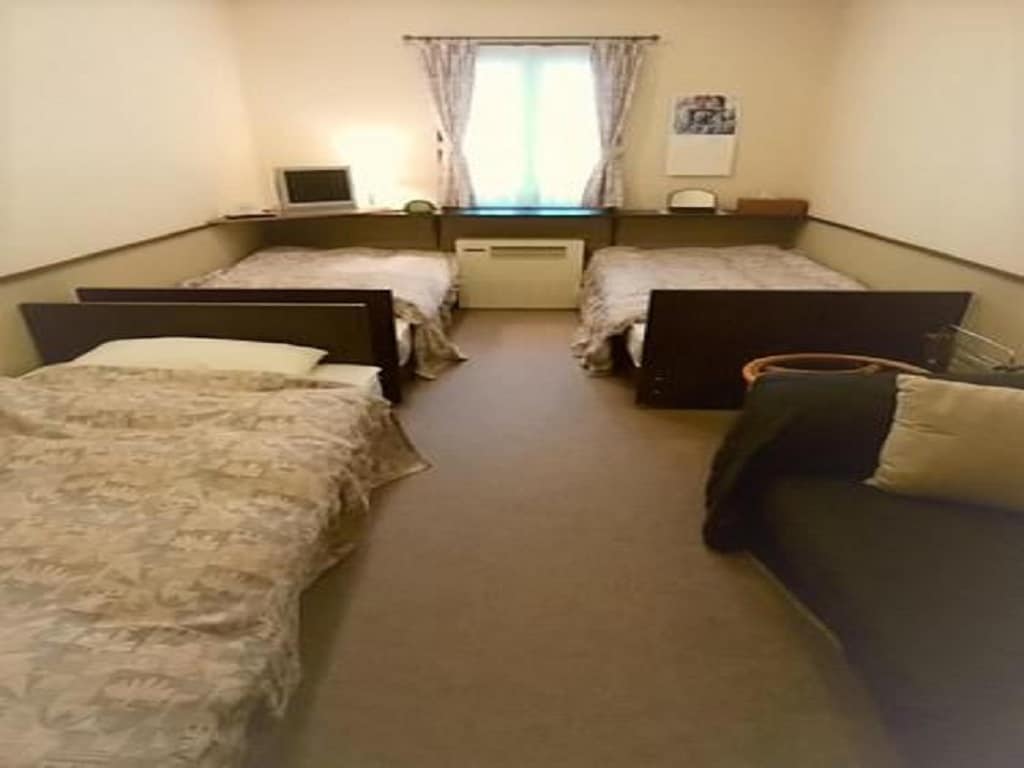 The room features three beds arranged in a spacious layout, with light-colored bedding. A window allows natural light to enter, and a small desk is positioned near the entrance. A seating area is included, providing additional comfort.