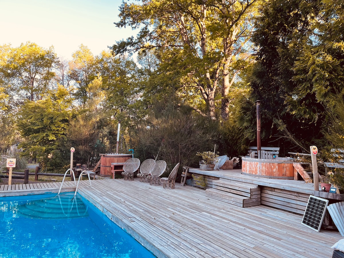 A wooden deck surrounds a clear swimming pool, featuring a set of four lounge chairs and a rustic seating area nearby. Lush greenery provides a natural backdrop, while a small solar panel is positioned on the deck.