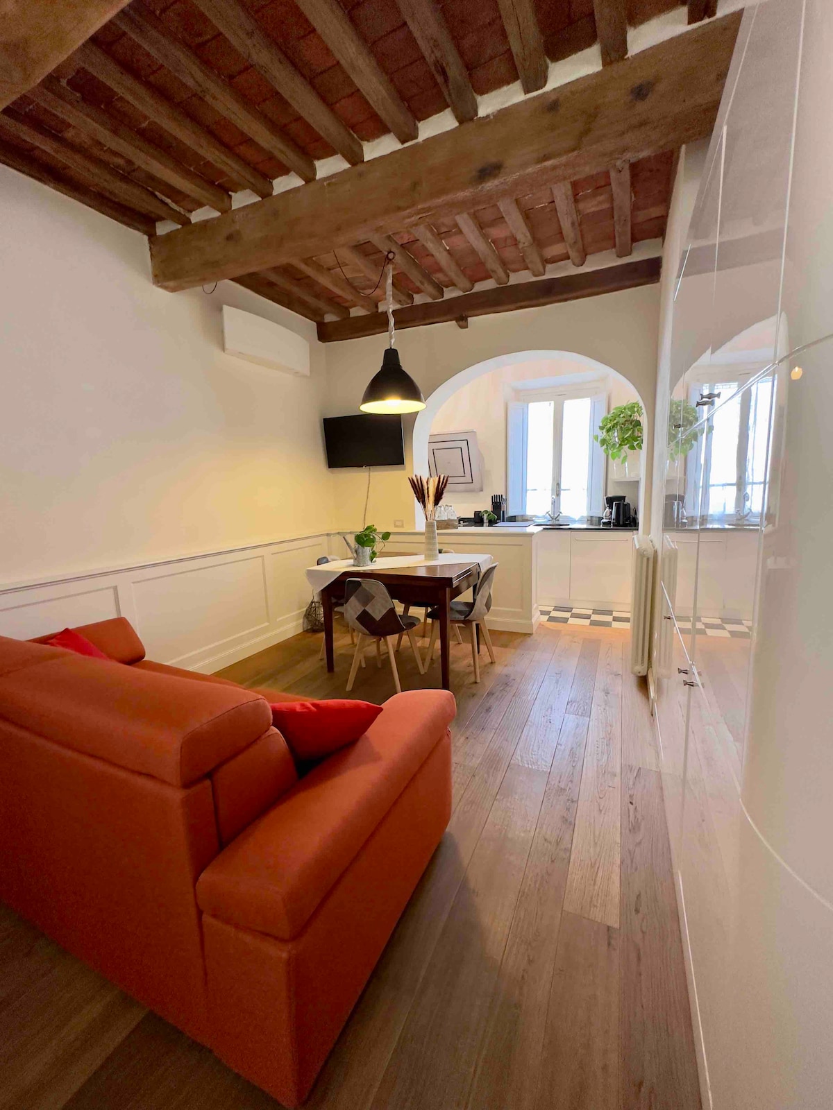 A cozy living space features a vibrant orange sofa positioned near a wooden dining table. The rustic wooden ceiling enhances the room's character. A modern television is mounted on the wall, and natural light streams through arched windows, illuminating the welcoming atmosphere.