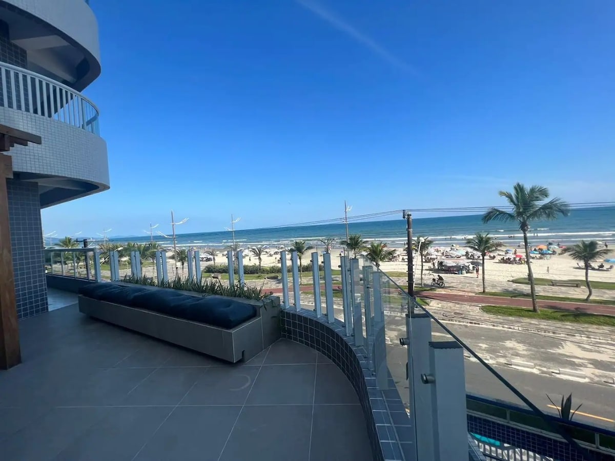 A spacious balcony offers a clear view of the beach and ocean. The outdoor area features a long seating space along the railing, with modern glass fencing enhancing the scenic perspective. Palm trees line the sandy shore, creating a relaxed coastal atmosphere.