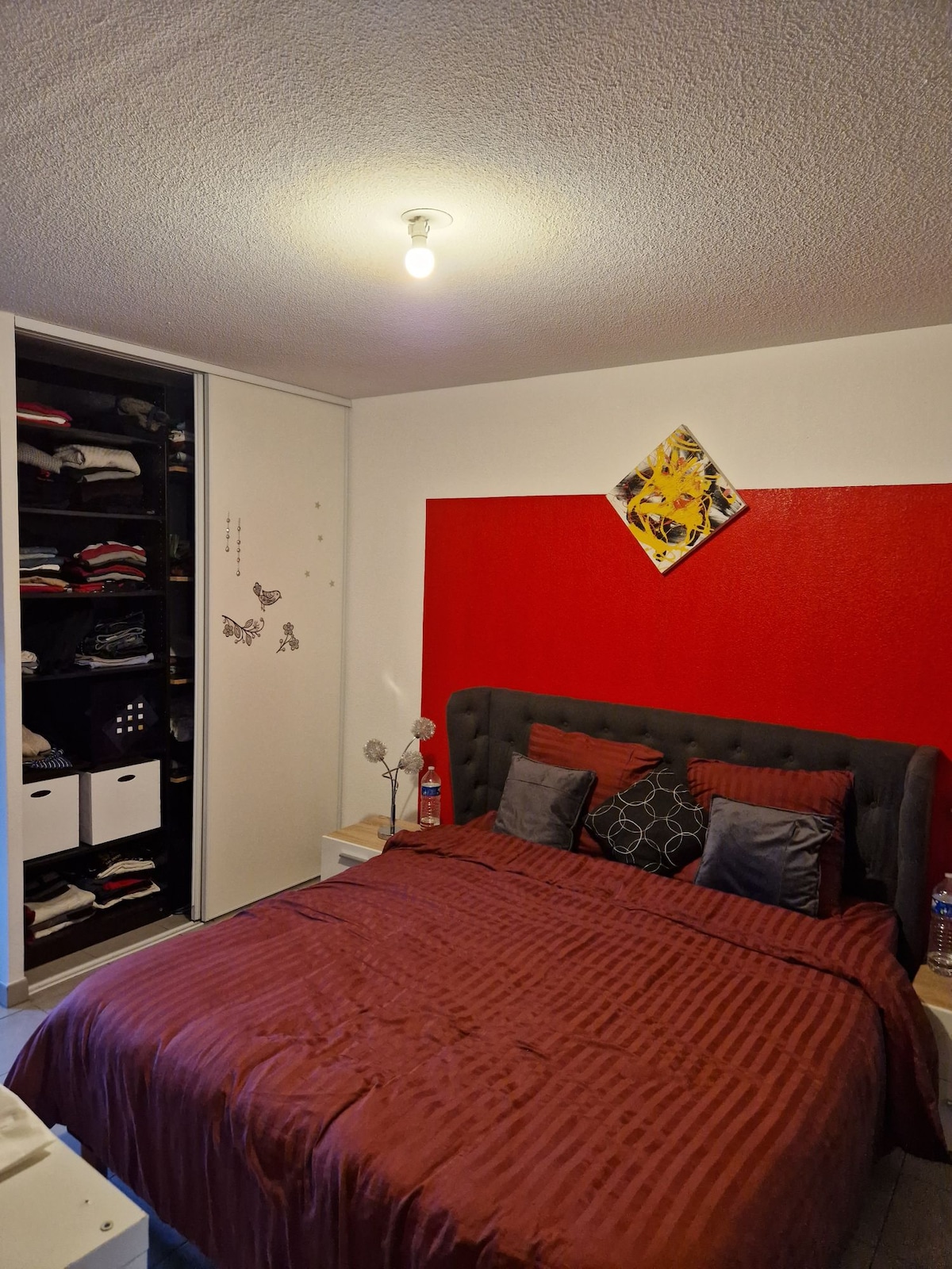 A cozy bedroom features a dark upholstered bed with accent pillows against a vibrant red wall. A wardrobe with an organized open section displays neatly folded clothes and decorative boxes. Soft overhead lighting provides a warm glow throughout the space.