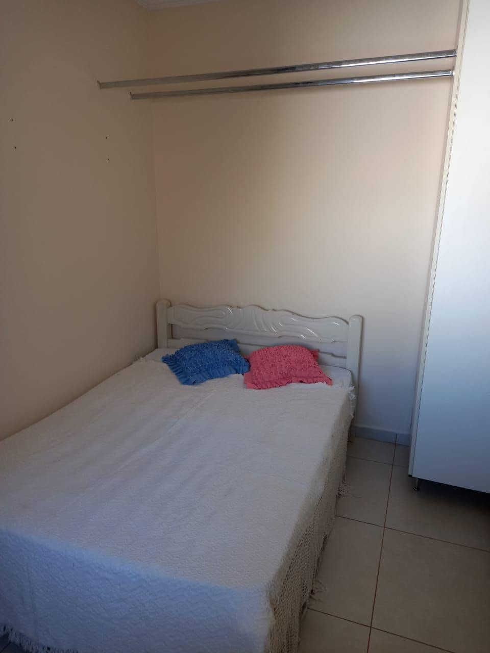 The room contains a spacious bed covered with a light-colored blanket. Two colorful throws are draped at the head of the bed. A simple closet is positioned against the adjacent wall, while the warm tones of the walls contribute to a calm environment.