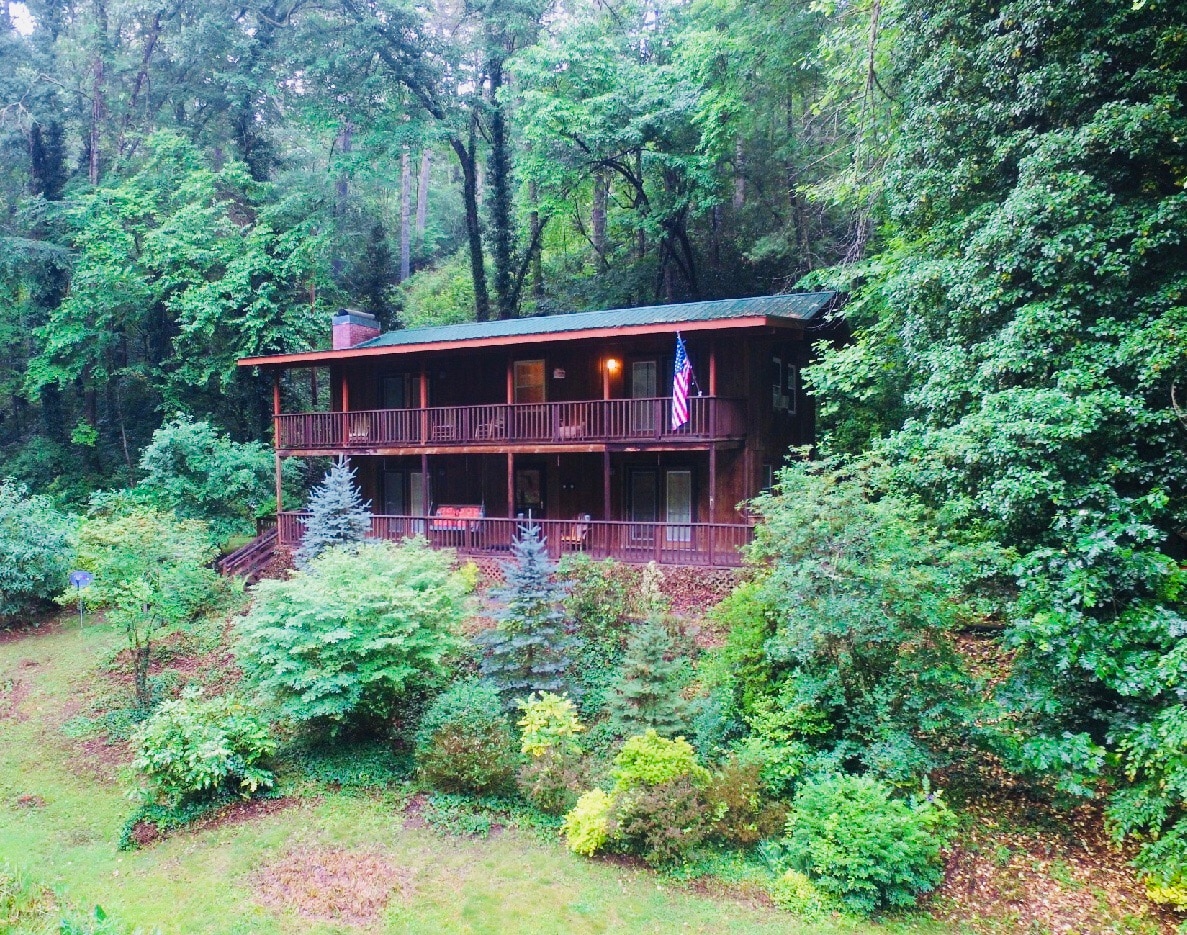 The large mountain home is surrounded by lush, mature trees, creating a serene natural setting. A spacious balcony overlooks the expansive yard, featuring several evergreen shrubs. The structure displays a dark wood exterior with multiple levels and an American flag gently waving in the breeze.