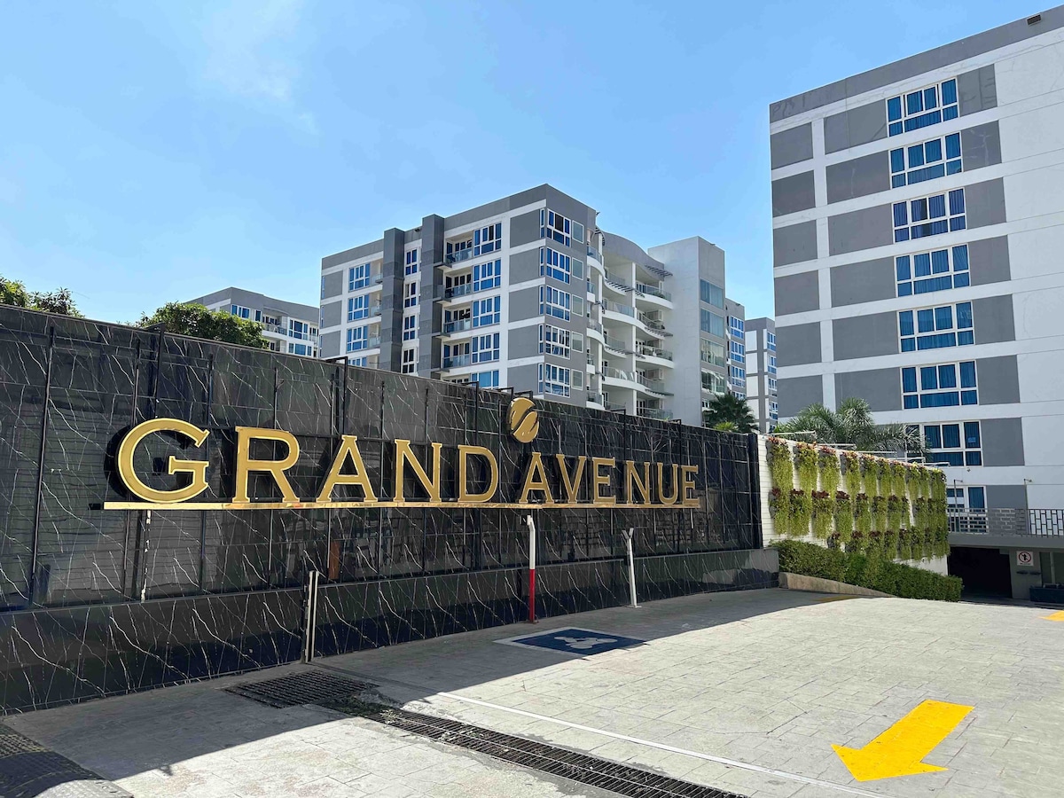 The Grand Avenue entrance features a prominent gold sign displaying the name 'GRAND AVENUE' against a dark stone backdrop. Surrounding foliage enhances the entrance area, while modern apartment buildings can be seen in the background under a clear blue sky.