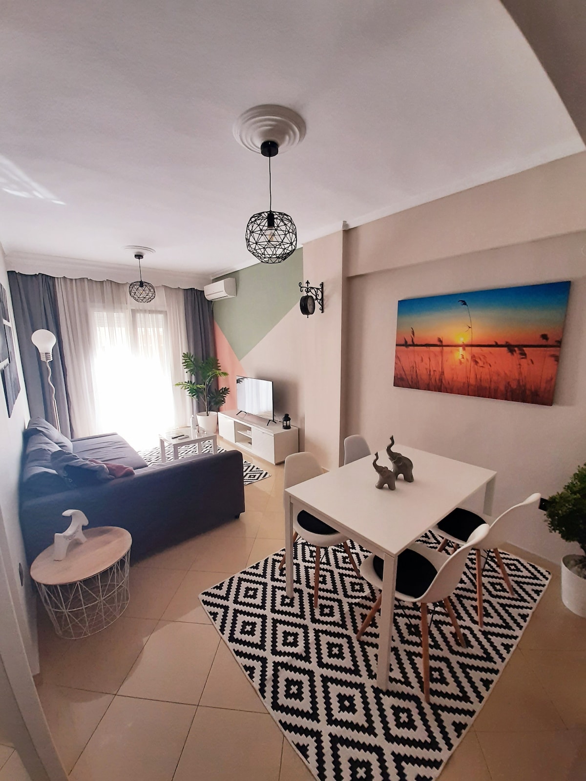A modern living space is showcased with a cozy sofa facing a wall-mounted television. A light-filled dining area features a white table surrounded by black and white chairs, complemented by a vibrant wall art piece. Natural light filters through sheer curtains.