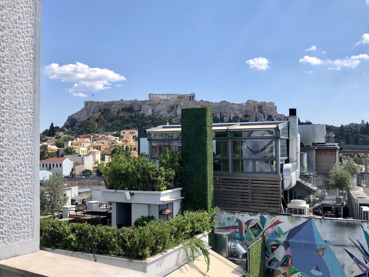 The image showcases a panoramic view of the Acropolis against a clear blue sky, with white clouds scattered above. In the foreground, a well-maintained terrace features greenery and seating areas, creating a welcoming outdoor space. The surrounding neighborhood displays a blend of modern and traditional architecture.
