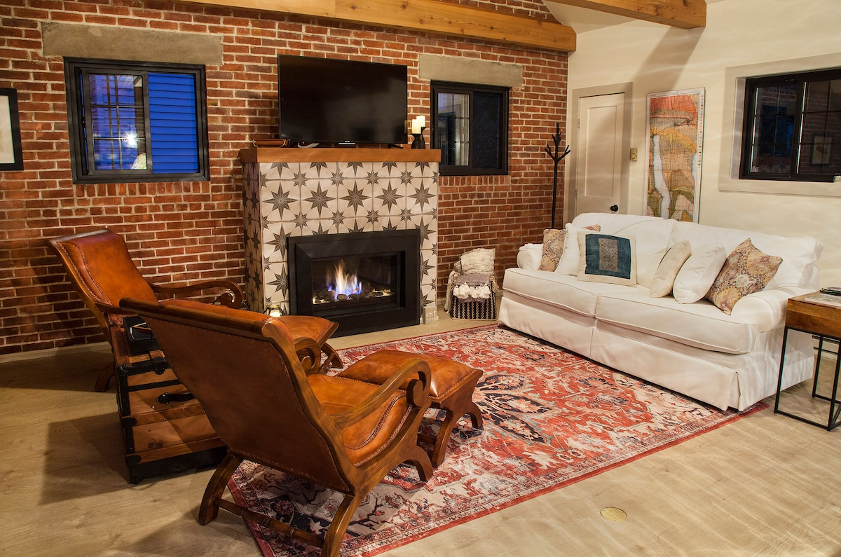 A cozy living area features exposed brick walls and a gas fireplace. A large white sofa is accompanied by matching armchairs on a patterned area rug. Natural light enters through the windows, with decorative art pieces adding character to the space.