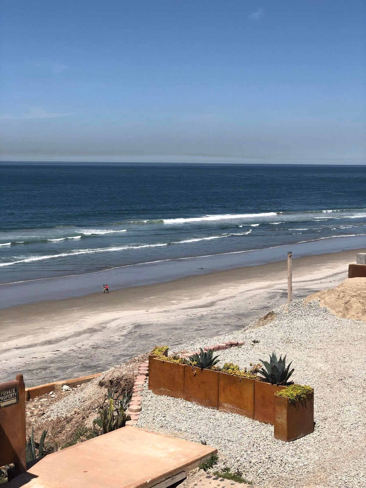 A sandy beach stretches before rolling waves under a clear blue sky. Gentle waves can be seen meeting the shore, while a few beachgoers enjoy the serene environment. A rocky and landscaped area is visible in the foreground, with low-growing plants and pebbles.