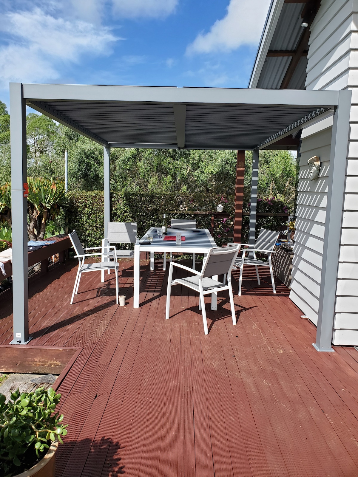 An outdoor patio area is presented, featuring a metal frame gazebo with a slatted roof. A glass-top table is surrounded by four white chairs. The deck is adorned with a potted plant, while lush greenery is visible in the background under a clear blue sky.
