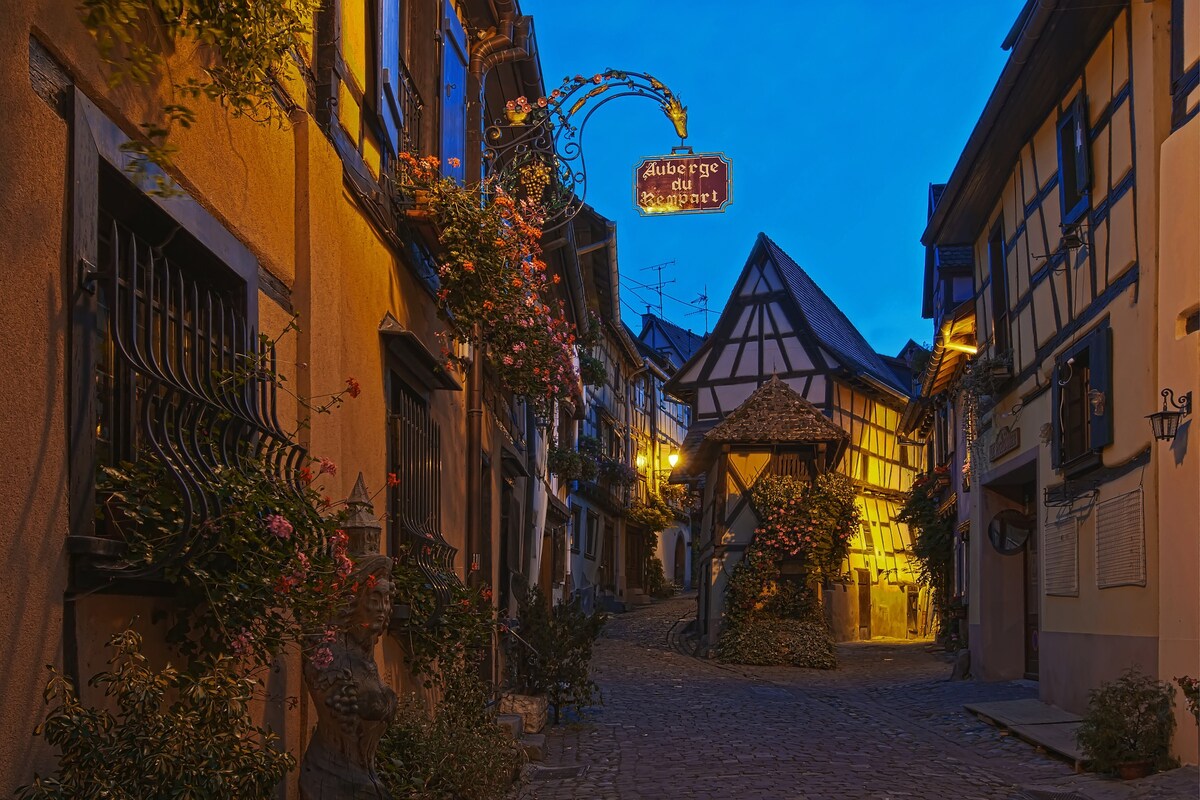A cobblestone street is illuminated by warm lights, revealing well-preserved half-timbered houses adorned with vibrant flowers. A decorative sign hangs above the narrow street, leading towards a quaint building at the end, framed by charming architecture typical of Alsace.