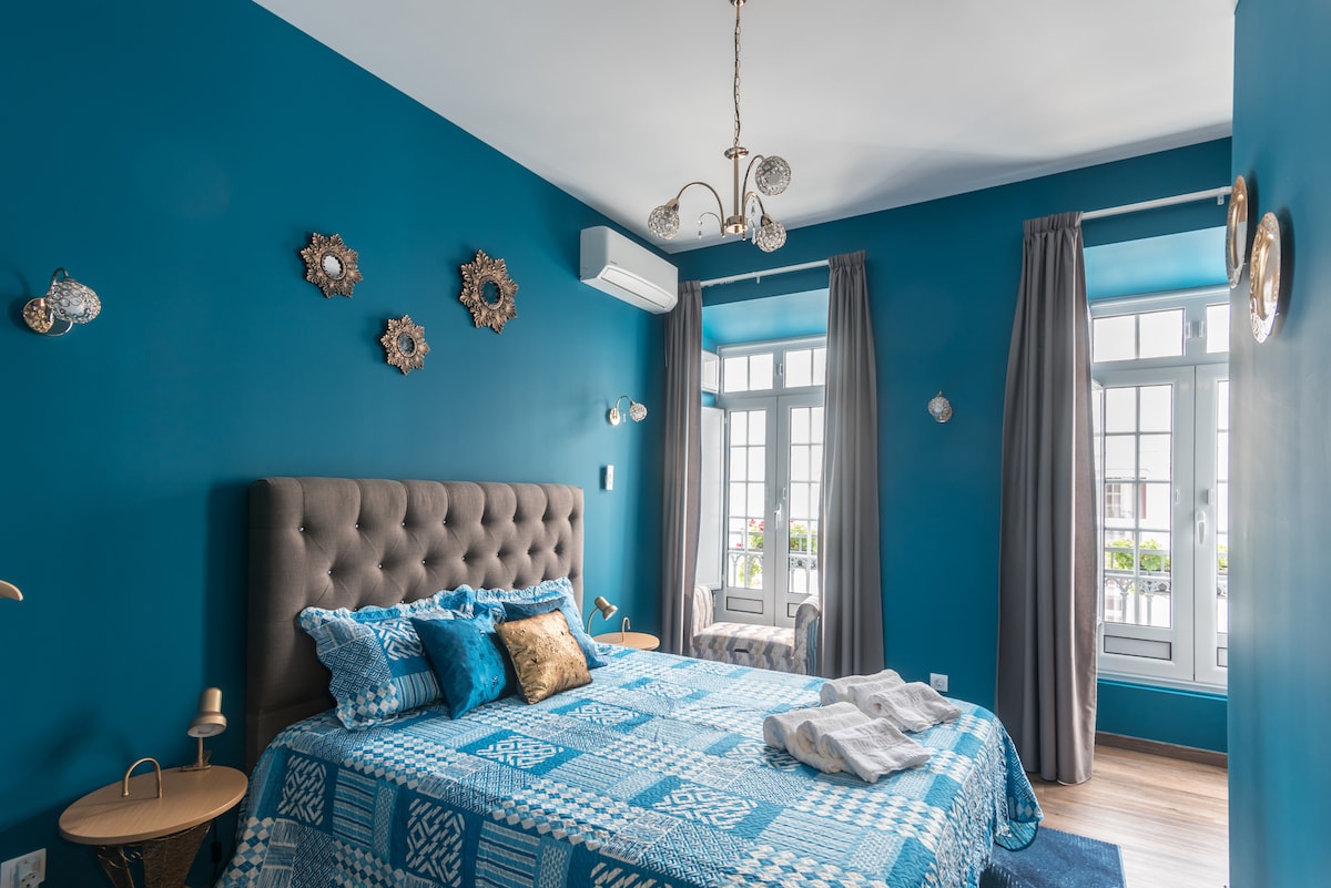 The bedroom features a deep blue accent wall, highlighting a plush bed adorned with a patterned blanket. Elegant light fixtures hang above, while soft curtains frame windows that allow natural light to brighten the space. Decorative wall art adds character to the room.