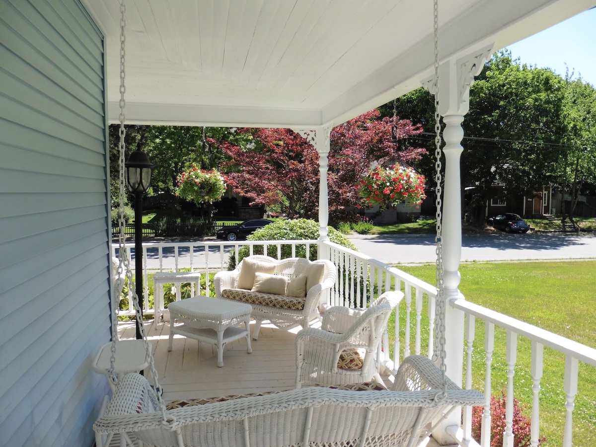 A charming front porch features wicker seating arrangements, including a loveseat and two armchairs, surrounding a small table. The white railings provide a view of the surrounding greenery, complemented by hanging flower baskets that add a touch of color to the space.