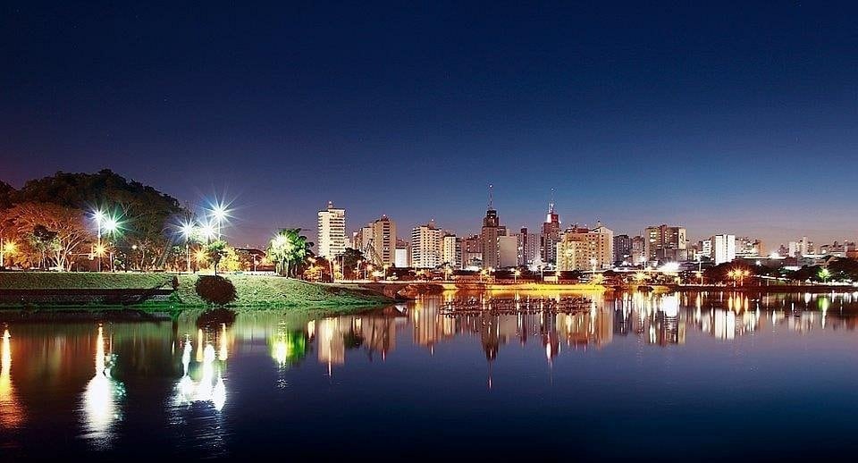 A panoramic view of a city skyline at dusk reflects in calm water. Bright lights from buildings and surrounding greenery illuminate the scene, showcasing a blend of nature and urban life. The tranquil atmosphere is enhanced by the serene lake reflecting the twinkling city lights.